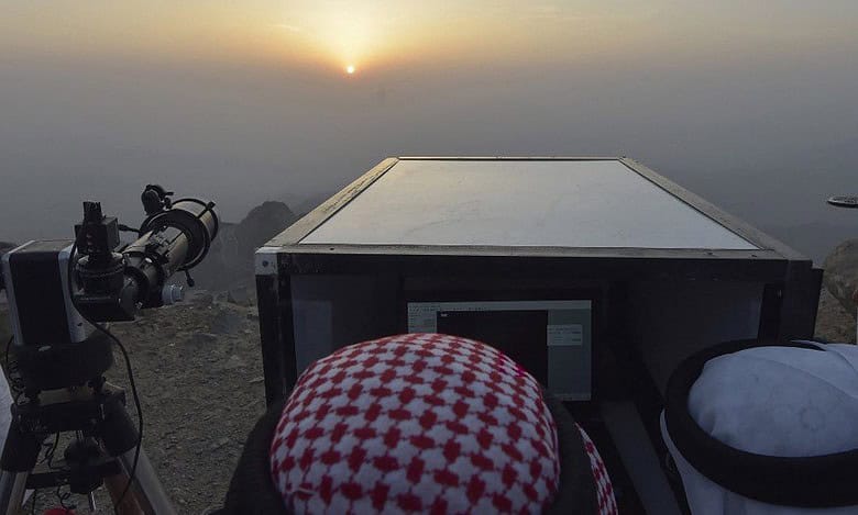 Moon sighting observation for Eid Al-Fitr at sunset, featuring astronomers in traditional Middle Eastern attire using a telescope and digital equipment on a mountaintop. The image captures the crescent moon sighting process, a crucial Islamic tradition for determining the start of Shawwal and Eid celebrations.