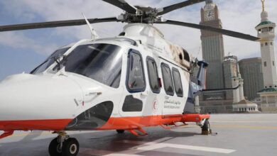 The image shows a Saudi Red Crescent Authority air ambulance helicopter parked on a helipad near the Grand Mosque in Makkah, Saudi Arabia. The helicopter is white with red and blue accents, and it features Arabic and English text indicating its affiliation with emergency medical services.
