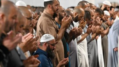 This image captures a group of Muslim worshippers standing in prayer at Makkah’s Grand Mosque during Isha and Taraweeh on the 29th night of Ramzan 1446 AH. The men, dressed in traditional attire such as thobes and kufis, are standing in rows with their hands raised in dua.