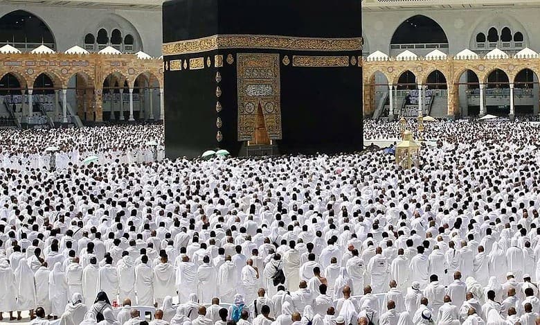 A vast congregation of Muslim worshippers, dressed in white ihram garments, gathers around the Kaaba at the Grand Mosque (Masjid al-Haram) in Makkah, Saudi Arabia, for the last Friday prayer of Ramzan. The sacred black-draped Kaaba stands at the centre, adorned with golden Arabic calligraphy.