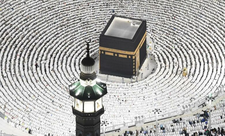 A crowd in white surrounds the Kaaba, a black cubic structure with gold details, in a lit courtyard at night. A green-lit minaret is in the foreground, during a religious gathering, likely Haj or Umrah in Mecca, Saudi Arabia.