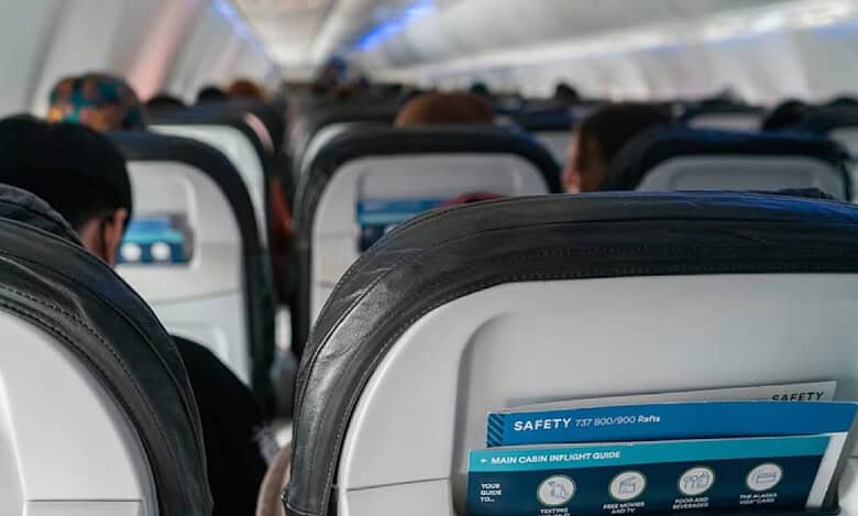 Interior view of an airplane cabin showing passengers seated with safety instruction cards visible in the seat pockets – economy class travel.