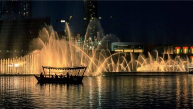 Traditional boat with people sailing on Burj Lake at night with Dubai Fountain water show in the background, lit with golden lights near Dubai Mall, UAE.