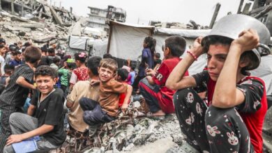 Children sit amid rubble in Gaza Strip, with worried expressions, in a crowded area of debris and destroyed buildings. A child wears a pot as a hat, reflecting distress.