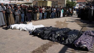 A large group of mourners stands in prayer over several bodies wrapped in black and white body bags, laid out on a paved ground in broad daylight.