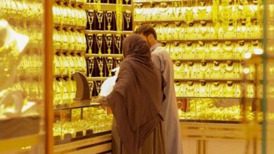A man and woman in traditional attire shopping for gold jewellery in a well-lit Dubai gold souk. The shop is filled with gold bangles, necklaces, and ornaments displayed in glass counters and wall panels, creating a luxurious, golden ambience.