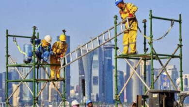 Construction workers on scaffolding at a building site in a Gulf city, wearing safety helmets and high-visibility clothing.