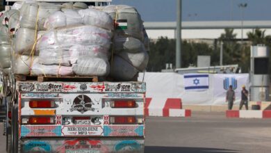 Aid truck loaded with supplies approaches Israeli checkpoint, with flags and guards in the background.