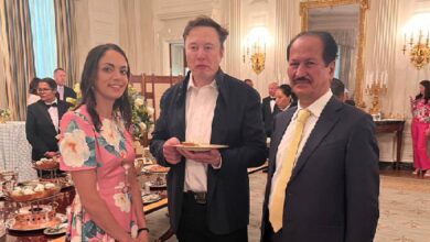 Hussain Sajwani stands with Elon Musk and Shivon Zilis at an elegant formal event. Musk holds a plate of food as they pose in a lavishly decorated room with chandeliers, a large mirror, and banquet tables.