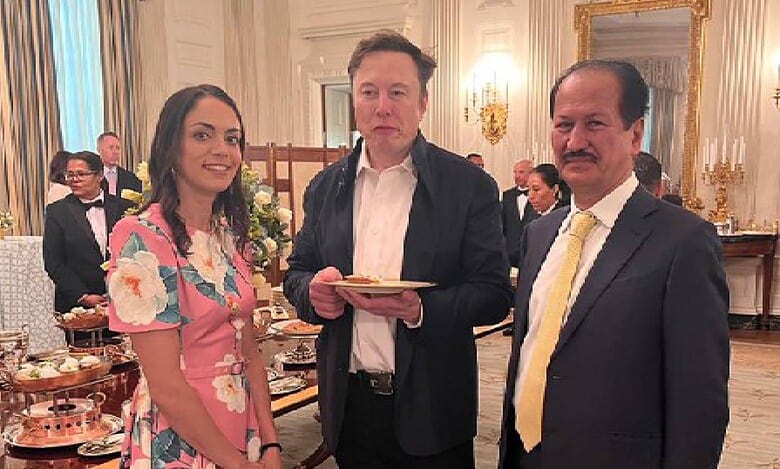 Hussain Sajwani stands with Elon Musk and Shivon Zilis at an elegant formal event. Musk holds a plate of food as they pose in a lavishly decorated room with chandeliers, a large mirror, and banquet tables.