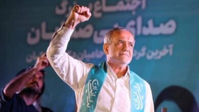 Photo of Iran Masoud Pezeshkian raising his fist during a campaign rally in Iran, wearing a turquoise sash with Persian writing, standing in front of a teal backdrop with Persian text.