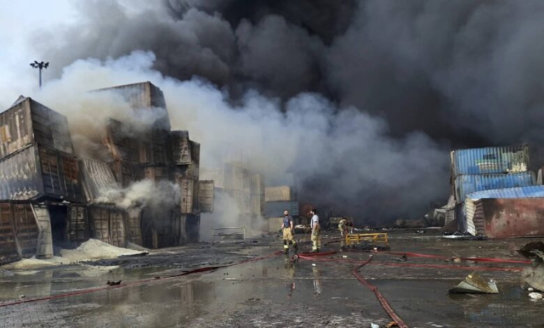 A building engulfed in flames, with thick black smoke billowing into the sky. Firefighters are present, working to extinguish the fire as debris and steam surround the factory.