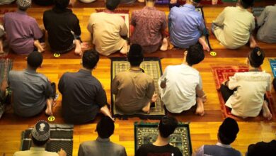 Group of men sitting on prayer mats in a mosque, preparing for congregational prayer. The image shows rows of worshippers from a top angle, wearing traditional and casual attire on a polished wooden floor."