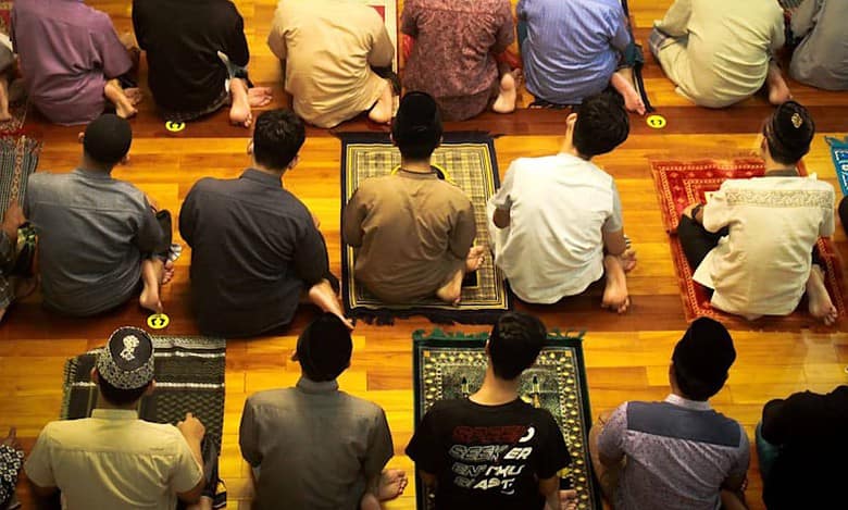 Group of men sitting on prayer mats in a mosque, preparing for congregational prayer. The image shows rows of worshippers from a top angle, wearing traditional and casual attire on a polished wooden floor."
