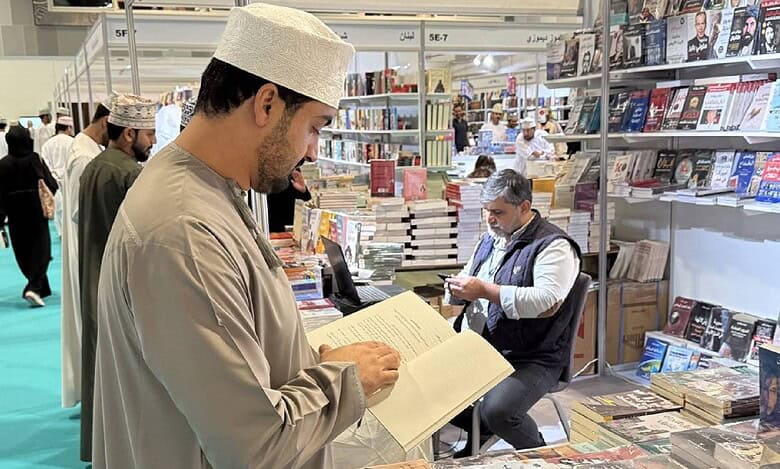 Visitor reading a book at a stall during the 29th Muscat International Book Fair, with shelves of Arabic books and exhibitors in the background.