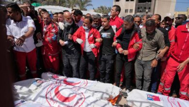A group of people, some in medical uniforms, stand in solidarity behind several bodies covered in Palestine Red Crescent shrouds during a public funeral.