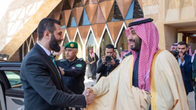 Saudi Crown Prince Mohammed bin Salman warmly shakes hands with Syrian interim President Ahmed al-Sharaa during a diplomatic visit to Riyadh, with security personnel and media present outside a modern government building.