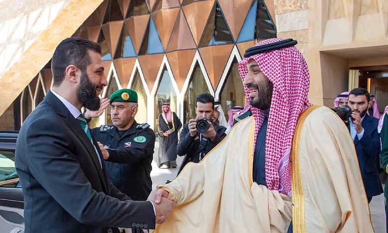 Saudi Crown Prince Mohammed bin Salman warmly shakes hands with Syrian interim President Ahmed al-Sharaa during a diplomatic visit to Riyadh, with security personnel and media present outside a modern government building.