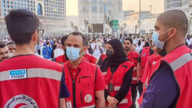 Saudi Red Crescent Authority volunteers in red vests and face masks coordinating efforts during Haj season in Makkah, Saudi Arabia, with pilgrims in the background.
