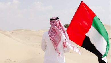 A man wearing traditional Emirati attire walks in the desert carrying the UAE national flag, under a partly cloudy sky.