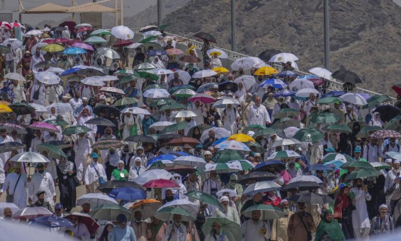 Large crowd of Haj pilgrims walking under umbrellas to protect themselves from the sun, during the annual pilgrimage in Saudi Arabia, with mountains in the background.