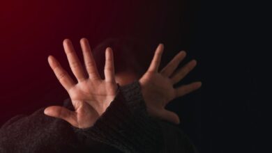 Person shielding their face with open hands in a defensive gesture, lit by red light against a dark background.