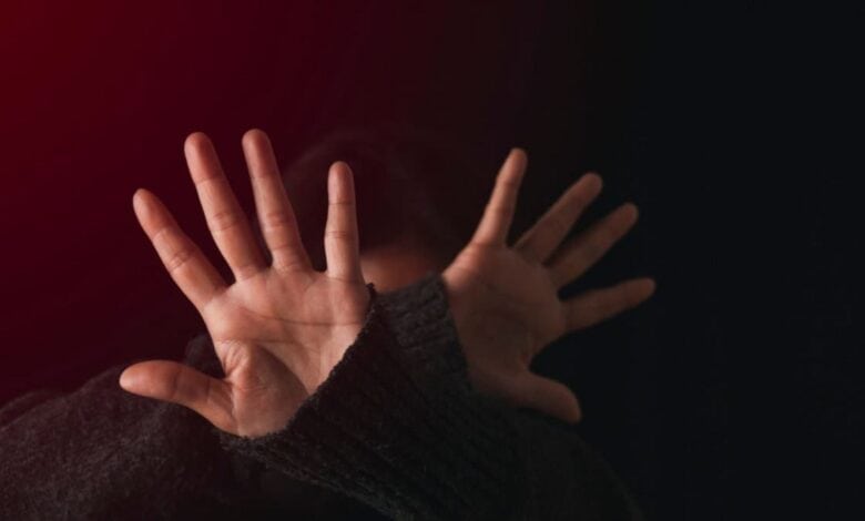 Person shielding their face with open hands in a defensive gesture, lit by red light against a dark background.