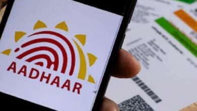Close-up of a smartphone showing the Aadhaar logo, with printed Aadhaar cards in the background on a wooden surface.