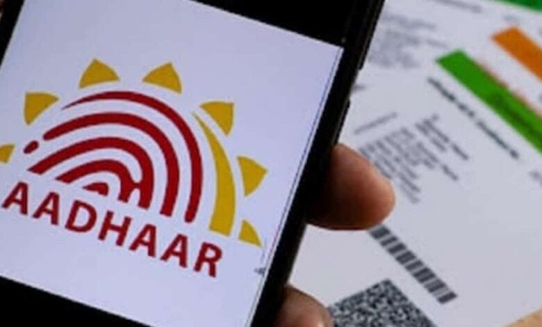 Close-up of a smartphone showing the Aadhaar logo, with printed Aadhaar cards in the background on a wooden surface.