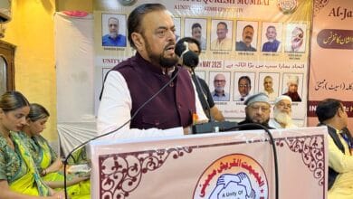 Man speaks at Al-Jamiatul Quresh Mumbai event, surrounded by guests and banners promoting unity in the Qureshi community.
