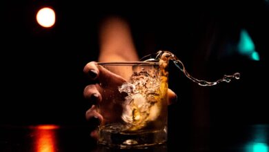A close-up of a hand holding a glass of alcoholic drink with ice, as the liquid splashes out dramatically against a dark background.
