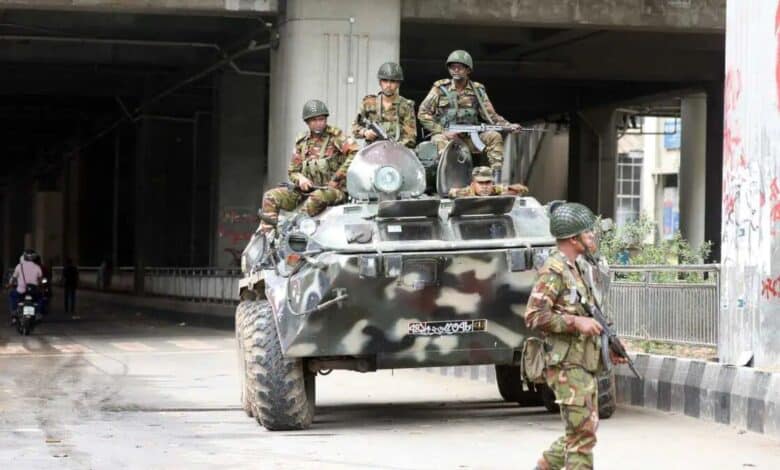 Soldiers in camouflage uniforms with rifles ride on an armored vehicle under a bridge, with one soldier patrolling on foot
