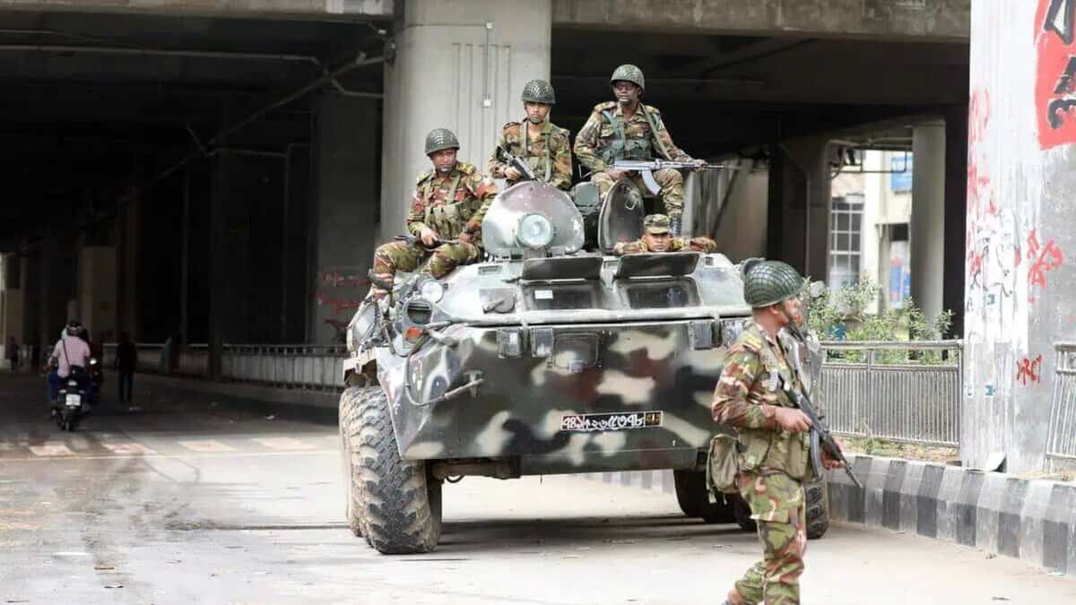 Soldiers in camouflage uniforms with rifles ride on an armored vehicle under a bridge, with one soldier patrolling on foot