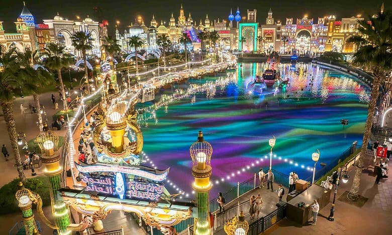 A vibrant night view of Dubai's Global Village featuring the Floating Market, colourful water show, and illuminated pavilions representing various countries.