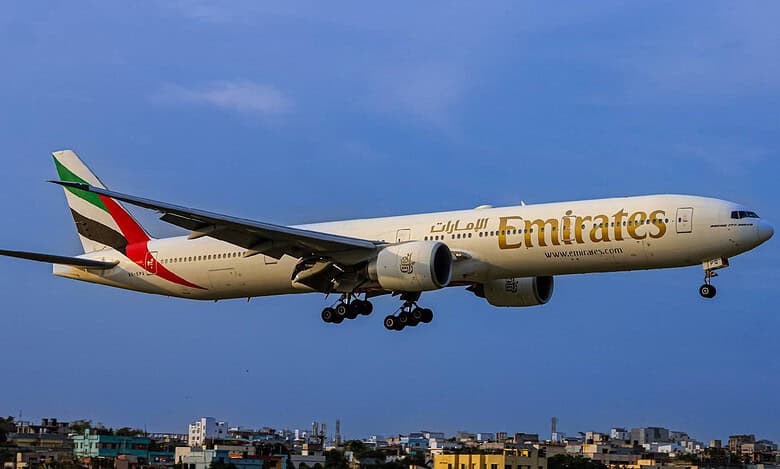 Emirates Boeing 777 aircraft flying low over a cityscape during landing, with landing gear deployed.