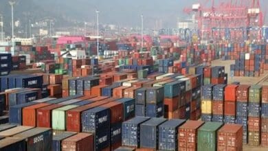 Hundreds of colorful shipping containers stacked at a busy port terminal with cranes and a hazy cityscape in the background