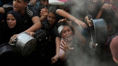 Crowd of desperate people in Khan Younis, Gaza, including children and adults, pressing against a barrier, holding out pots and pans for food aid.