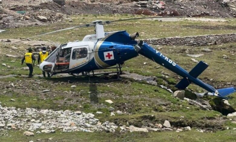 A medical helicopter with a red cross, crashed on a grassy, rocky terrain in Kedarnath. Two rescuers in yellow vests assist near the wreckage.