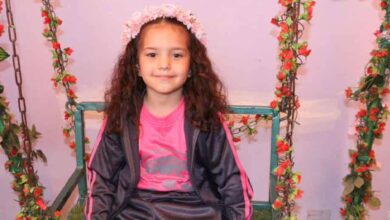 A smiling young Palestinian girl, Hind Rajab, wearing a pink sports outfit and floral headband, sits on a decorated swing surrounded by red flowers. Her tragic death in Gaza has drawn global attention and led to an ICC war crimes complaint.