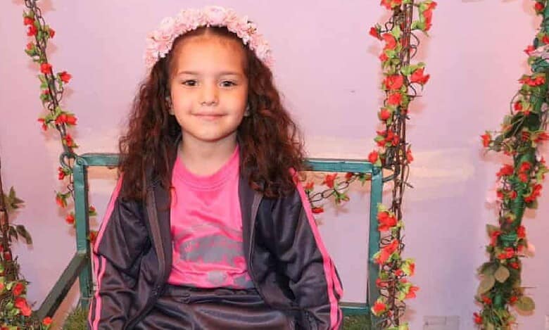 A smiling young Palestinian girl, Hind Rajab, wearing a pink sports outfit and floral headband, sits on a decorated swing surrounded by red flowers. Her tragic death in Gaza has drawn global attention and led to an ICC war crimes complaint.