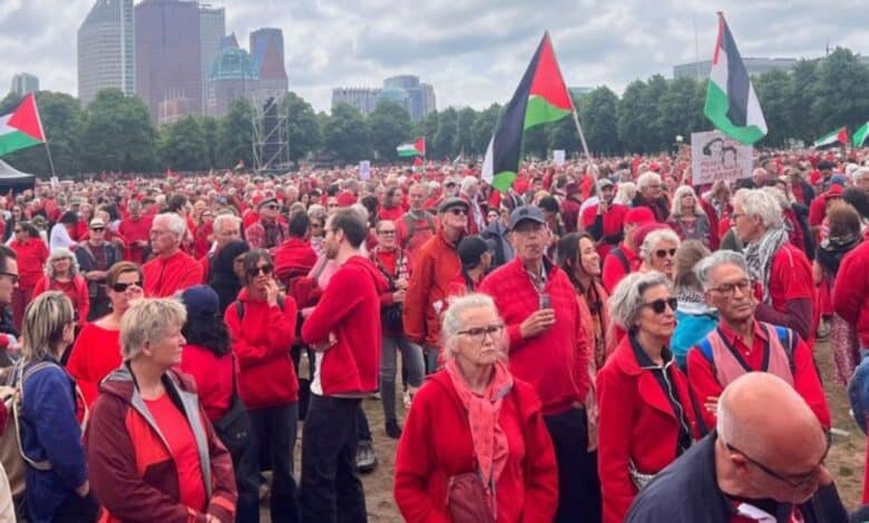 Large crowd dressed in red gathers at a protest with Palestinian flags and signs calling for an end to genocide, under cloudy skies.