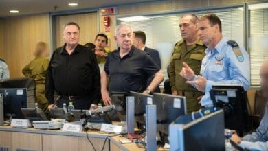 A group of officials in a control room with computers, discussing strategy. Some wear military uniforms, others casual attire, in a serious meeting setting.