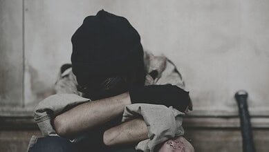 A distressed person sits curled up on the floor with head buried in arms, wearing a black beanie and torn clothing, suggesting emotional trauma or abuse.