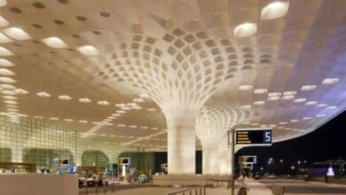 A spacious airport terminal with a modern ceiling design featuring illuminated, tree-like pillars and a perforated pattern. A digital sign displays gate numbers.
