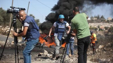 Journalists wearing press vests and helmets reporting near burning barricades during clashes in Palestine, with smoke billowing in the background.