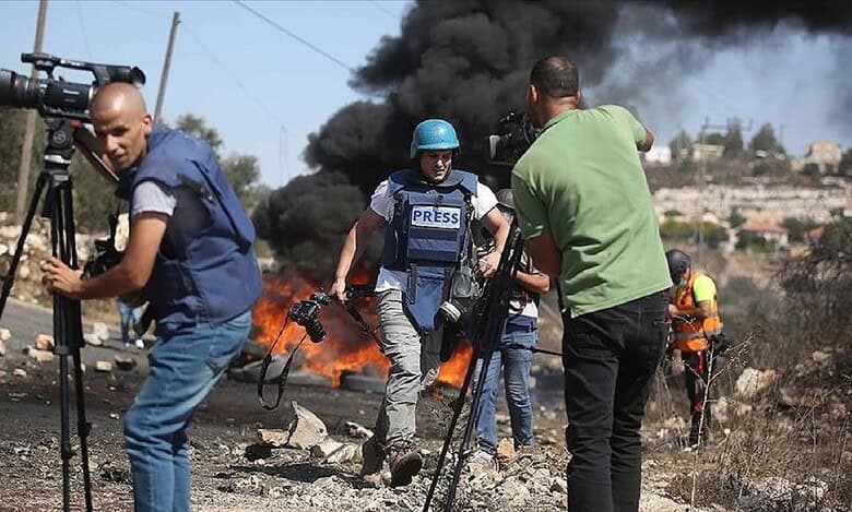 Journalists wearing press vests and helmets reporting near burning barricades during clashes in Palestine, with smoke billowing in the background.
