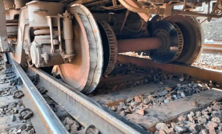 Close-up of a train's undercarriage, showing wheels on tracks with gravel ballast. The mechanical components, including axles and suspension, are visible.