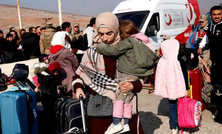 A mother carrying her child walks with luggage amid a crowd of refugees and aid workers near a Turkish Red Crescent van.