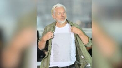 The image displays a picture of WikiLeaks founder Julian Assange at the 78th Cannes Film Festival in France wearing a T-shirt with the names of Palestinian children killed by Israeli forces since 2023.