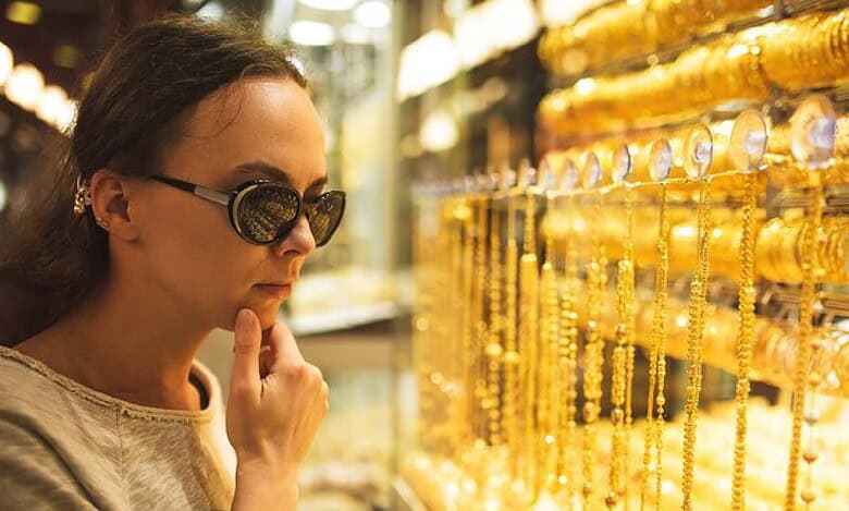 A woman wearing sunglasses closely examines gold jewellery displayed in a well-lit shop in Dubai.